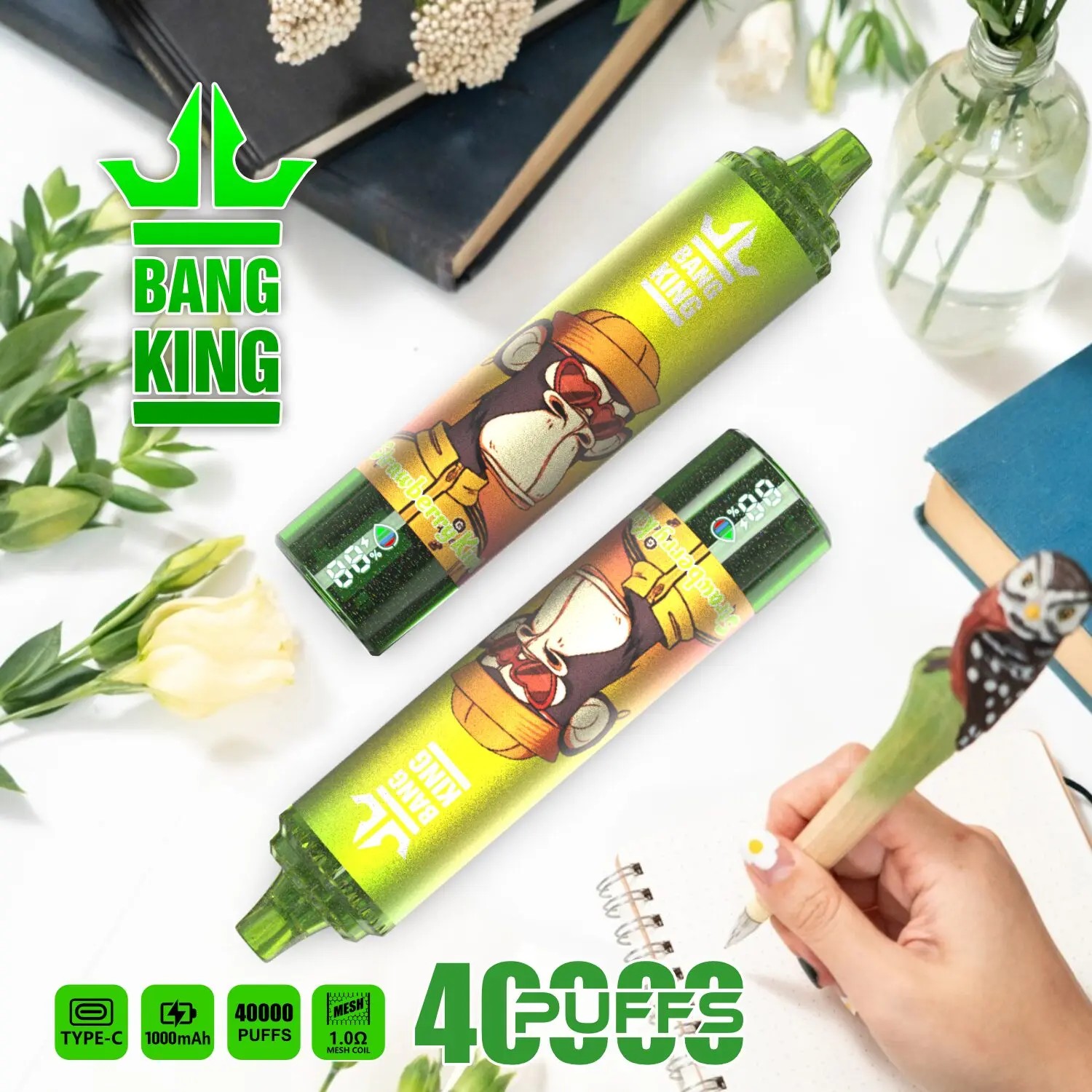 Two green Bang King disposable vape devices with a cartoon gorilla wearing a hat on the label, surrounded by green plants, flowers, books, and a hand holding a pen with an owl figure, on a white surface.