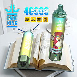 Two colorful Bang King disposable vape devices with a monkey illustration, placed on top of an open book with text in an Asian language, next to a pair of glasses and a white cup on a saucer.